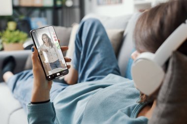 Evdeki kanepede uzanmış akıllı telefonuyla sosyal medya videoları izleyen bir kadın.