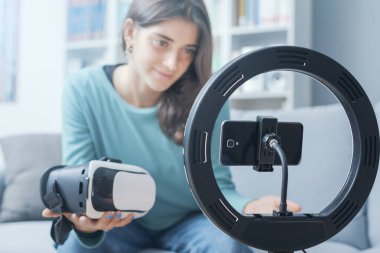 Sosyal medya profilleri için videolar yapan mutlu bir kız elinde bir VR kulaklık tutuyor ve akıllı telefon ekranına konuşuyor.