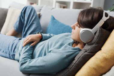 Kulaklık takan ve kanepede uzanan genç bir kadın dinleniyor ve müzik dinliyor.