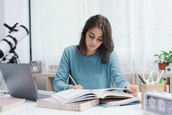 Smart student sitting at desk at home and doing homework, she is writing down notes on a notebook