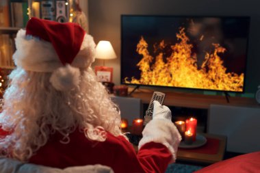 Noel Baba kanepede dinleniyor ve TV 'de yanan bir şömine izliyor. Elinde uzaktan kumanda var.