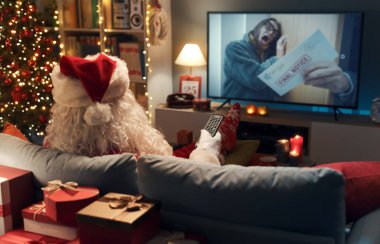 Mutlu Noel Baba evde kanepede dinleniyor ve TV 'de film izliyor.