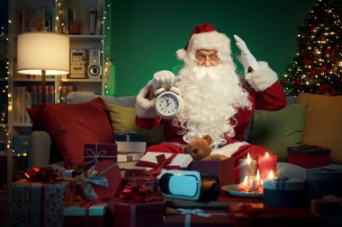 Stressed Santa Claus preparing gifts for Christmas at home and holding the alarm clock, he is late for the delivery clipart