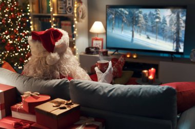 Happy Santa Claus relaxing on the couch at home and watching movies on TV clipart