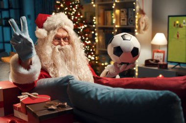 Mutlu Noel Baba kanepede oturup TV 'de futbol seyrediyor. Elinde bir top tutuyor ve Ziyaretçi tabelası yapıyor.