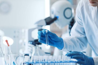 Android yapay zeka robotu laboratuarda mikroskop kullanıyor, bir kadın ön planda şişeler tutuyor.