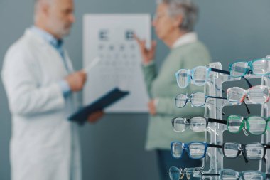 Senior woman having an eye test and talking with her eye care specialist, collection of eyeglasses in the foreground clipart