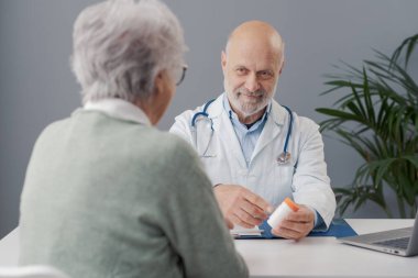 Doktor ofisinde çalışıyor, yaşlı bir kadına reçeteli ilaç veriyor ve gülümsüyor.