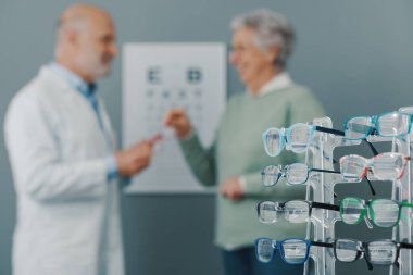 Kıdemli kadın göz testi yaptırıyor ve göz bakımı uzmanıyla konuşuyor. Ön planda gözlük koleksiyonu var.