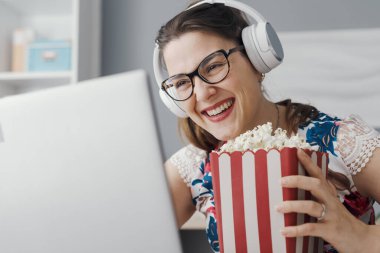 Happy woman sitting at her desk at home, she is watching online movies on the laptop and eating popcorn clipart