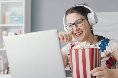 Happy woman sitting at her desk at home, she is watching online movies on the laptop and eating popcorn clipart