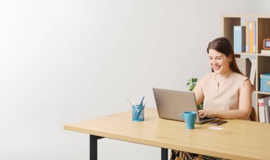 Masasında oturmuş dizüstü bilgisayarında komik videolar izleyen neşeli bir kadın gülümsüyor ve gülüyor.