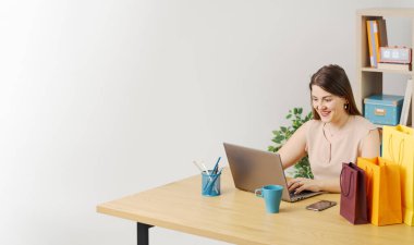Masada oturan mutlu kadın dizüstü bilgisayarıyla, iş ve ticaret konseptiyle online alışveriş yapıyor.