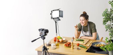 Gülümseyen profesyonel youtuber yemek pişirme videosu çekiyor, kameraya konuşuyor.