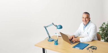 Ofisinde poz veren kendine güvenen bir doktor, masada oturuyor, dizüstü bilgisayarına yazıyor ve kameraya gülümsüyor.