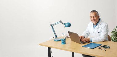 Confident doctor posing in his office, he is sitting at the desk, typing on a laptop and smiling at camera clipart