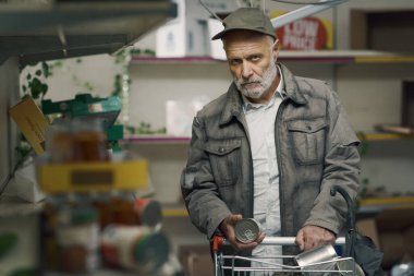 Desperate senior man pushing a full trolley in a destroyed supermarket: food shortage, war and hunger concept clipart