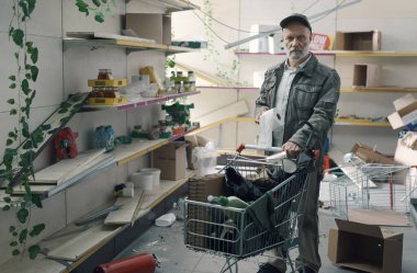 Mahvolmuş bir süpermarkette market alışverişi yapan üzgün adam, elinde tuvalet kağıdı tutuyor.