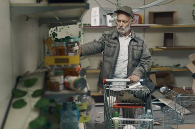 Desperate senior man searching for food in a post-apocalyptic destroyed supermarket with empty broken shelves: hunger, crisis, war and catastrophes concept clipart