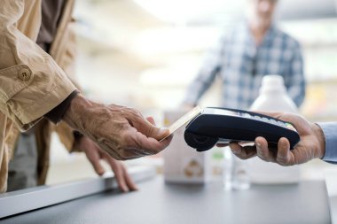Dükkandan alışveriş yapan yaşlı adam, kredi kartıyla ödeme yapıyor, elleri kapalı.