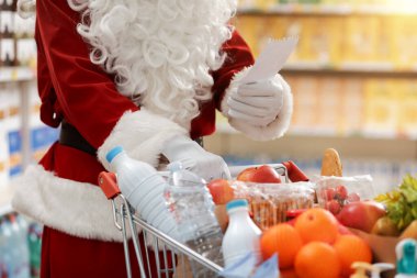 Noel Baba markette market alışverişi yapıyor, alışveriş arabasını itiyor ve alışveriş listesini kontrol ediyor, eller kapalı.