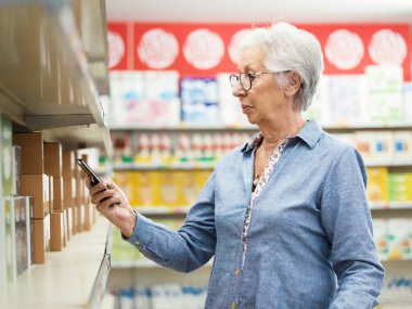 Üst düzey kadın süpermarketten alışveriş yapıyor, gıda tarayıcı uygulaması kullanarak raftaki ürünleri kontrol ediyor.