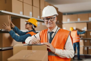 İşteki gülümseyen depo müdürü, bir belge yazıyor ve siparişleri kontrol ediyor: lojistik ve teslimat hizmeti kavramı