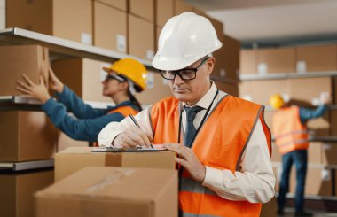 İş yerindeki profesyonel depo müdürü, bir belge yazıyor ve siparişleri kontrol ediyor: lojistik ve teslimat hizmeti kavramı