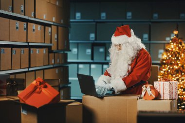 Noel Baba depoda dizüstü bilgisayarla çalışıyor. Teslimat kutularını daktilo edip hazırlıyor.