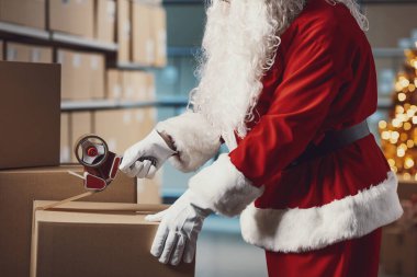 Noel Baba dağıtım deposunda karton bir teslimat kutusunu bantlıyor, Noel ve tatil konsepti.