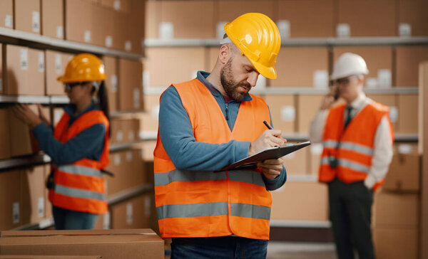 Professional warehouse worker posing with a safety helmet and holding a clipboard, logistics and shipment concept