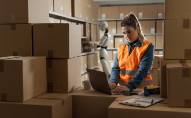 Depo işçisi ve yapay zeka robotu birlikte çalışıyor: kadın dizüstü bilgisayarda yazıyor ve robot kutuları taşıyor