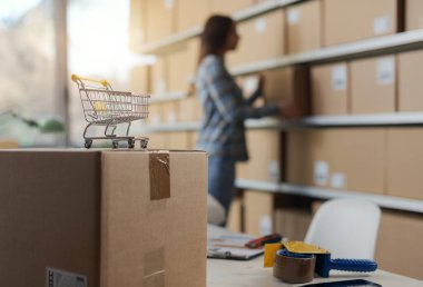 E-ticaret şirketi sahibi depoda teslimat kutuları taşıyor, ön planda küçük alışveriş arabası: çevrimiçi alışveriş ve teslimat kavramı