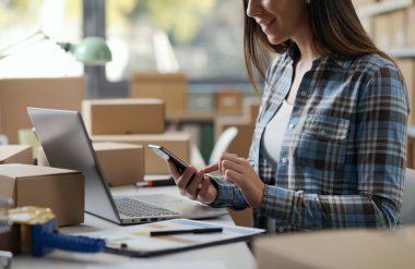 E-ticaret işyeri sahibi tezgahta oturuyor ve akıllı telefon kullanıyor.