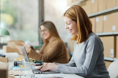 Depoda çalışan gülümseyen e-ticaret çalışanları, bir dizüstü bilgisayarda yazıyorlar ve malları teslimat kutularına koyuyorlar.