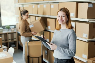 Depoda çalışan E-ticaret çalışanları teslimat kutularını taşıyorlar ve siparişleri kontrol ediyorlar. Bir kadın panoya yazı yazıyor.