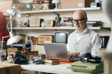 İnternet üzerinden antika nesneler satan kıdemli bir işyeri sahibi, mağazasındaki bir dizüstü bilgisayara bağlanıyor, e-ticaret konsepti.