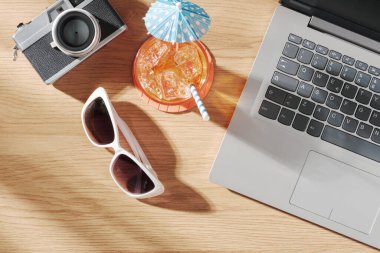 Masadaki dizüstü bilgisayar, kamera, kokteyl ve güneş gözlükleri: yaz tatilleri ve rahatlama konsepti, üst görünüm