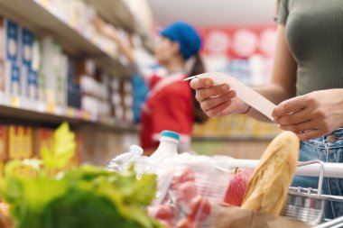 Kadın market alışverişi yapıyor ve makbuzu kontrol ediyor, eller kapalı.