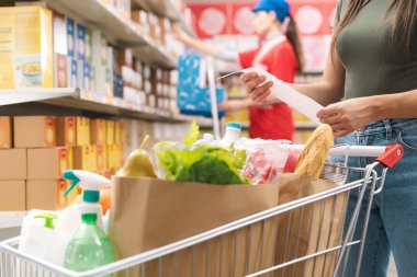 Kadın market alışverişi yapıyor ve makbuzu kontrol ediyor, eller kapalı.