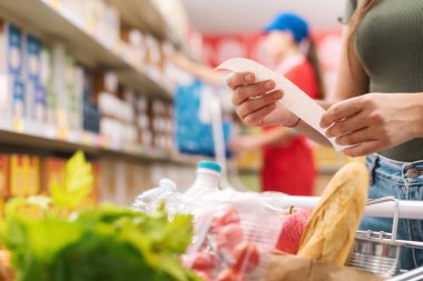 Kadın market alışverişi yapıyor ve makbuzu kontrol ediyor, eller kapalı.