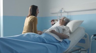 Hasta annesini ziyarete gelen genç bir kadın hastane yatağında yatıyor, birlikte konuşuyor ve gülümsüyorlar.