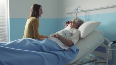 Hasta annesini ziyarete gelen genç bir kadın hastane yatağında yatıyor, birlikte konuşuyor ve gülümsüyorlar.