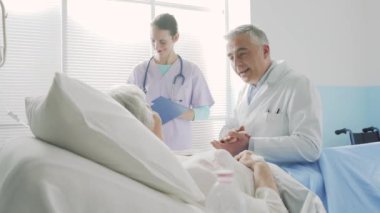 Kıdemli bir hastayla konuşan ve onu destekleyen bir hemşire gülümsüyor ve yanında duruyor.