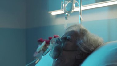 Hasta annesine yardım eden genç bir kadın, hastane yatağında oksijen maskesiyle uyuyor.