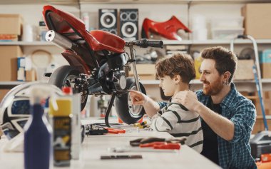 Gururlu bir baba çocuğuna garajda motosiklet kullanmayı öğretiyor. Gülümsüyor ve işaret ediyor.