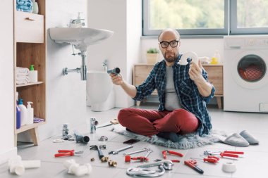 Kafası karışmış, banyo lavabosunu tek başına tamir etmeye çalışan kafası karışık bir adam. Etrafı aletlerle çevrili ve kanalizasyon parçalarını tanıyamıyor.