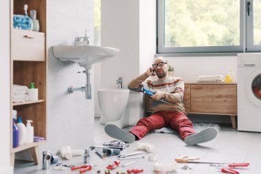 Hüsrana uğramış bir adam banyo lavabosunu tek başına tamir edemiyor yerde oturuyor ve akıllı telefonuyla profesyonel bir tesisatçıyı arıyor.