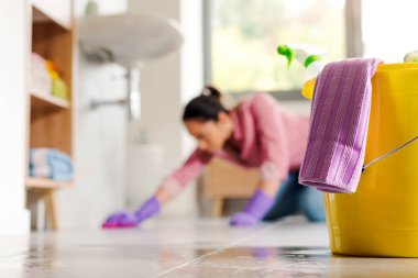 Genç bir kadın banyo zemininde diz çöküyor ve ön planda plastik bir kovayla bez parçaları temizliyor.