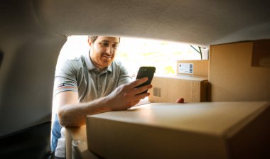 Teslimatçı bir minibüsü yüklüyor ve akıllı telefonunda uygulamalar kullanıyor.